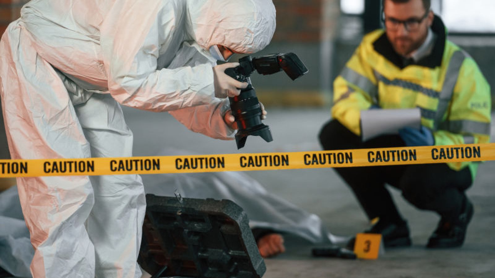 Forensic expert in a hazmat suit photographing a crime scene with police oversight.